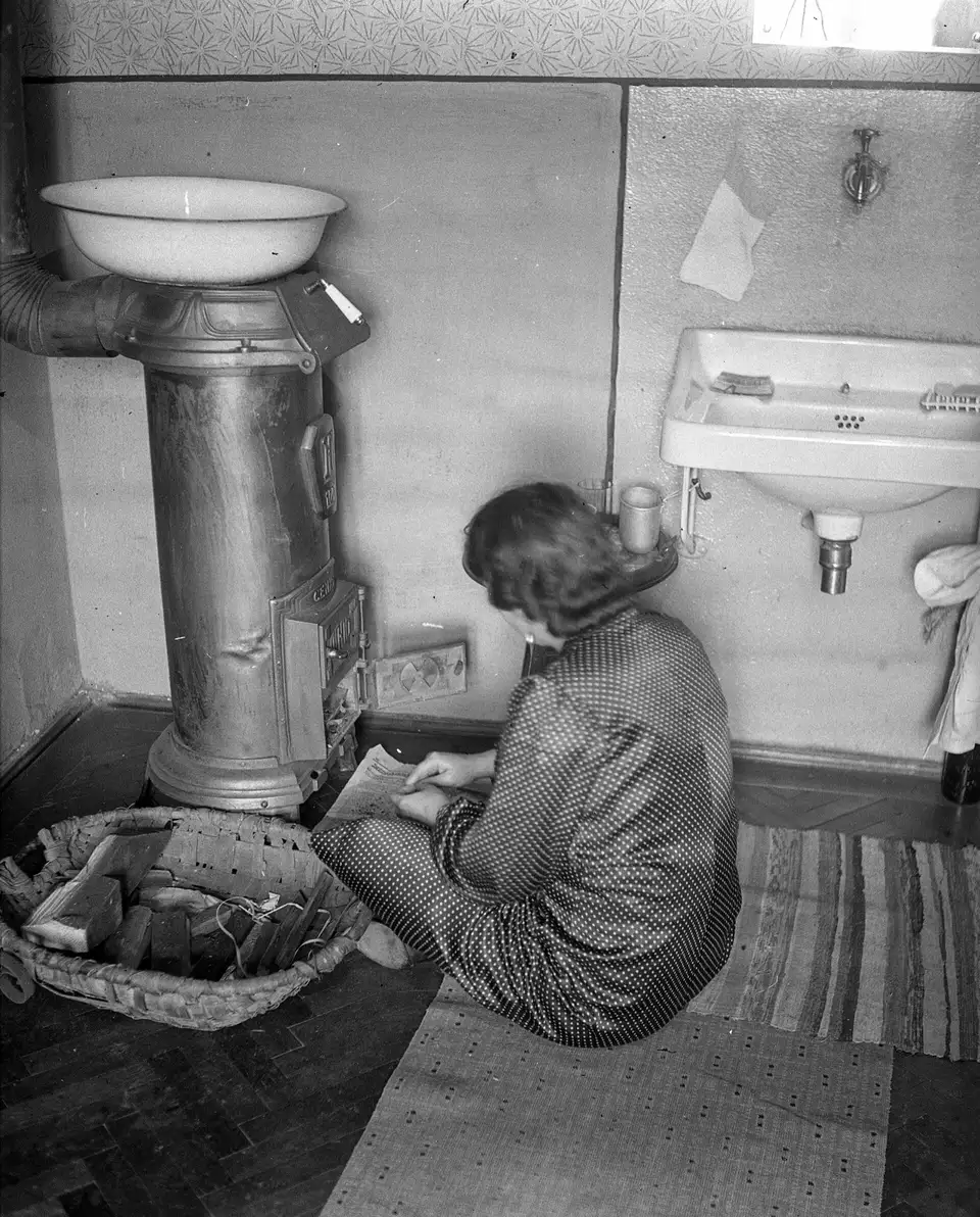 Kobieta kuca przy żeliwnym piecyku w łazience i sięga do wiklinowego kosza z drewnem